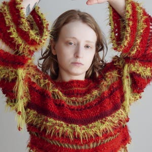 May include: A long-sleeved, cropped sweater featuring horizontal stripes in shades of deep red, burgundy, and bright yellow. The garment is constructed with a textured, fuzzy yarn that creates a shaggy, layered appearance throughout the body and sleeves.