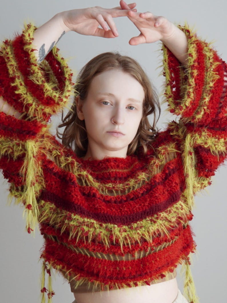 May include: A long-sleeved, cropped sweater featuring horizontal stripes in shades of deep red, burgundy, and bright yellow. The garment is constructed with a textured, fuzzy yarn that creates a shaggy, layered appearance throughout the body and sleeves.