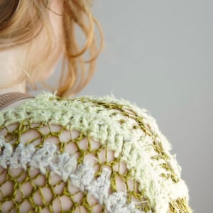 May include: A crochet top featuring an open-knit mesh pattern in shades of olive green, sage, and cream. The garment is constructed with alternating rows of textured, fuzzy yarn and a thinner, smooth yarn, creating a layered, dimensional appearance.