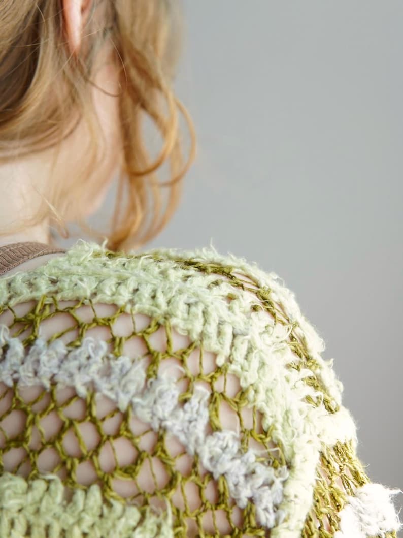 May include: A crochet top featuring an open-knit mesh pattern in shades of olive green, sage, and cream. The garment is constructed with alternating rows of textured, fuzzy yarn and a thinner, smooth yarn, creating a layered, dimensional appearance.