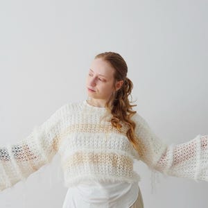 May include: A cropped, long-sleeved jumper featuring a loose, open-knit texture with alternating horizontal stripes of cream and soft beige. The garment includes frayed, fuzzy yarn edges along the hem and cuffs, creating a textured appearance.