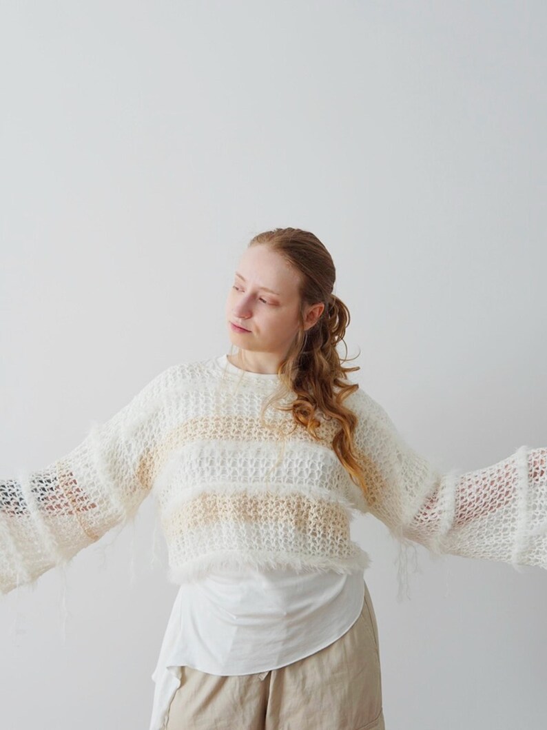 May include: A cropped, long-sleeved jumper featuring a loose, open-knit texture with alternating horizontal stripes of cream and soft beige. The garment includes frayed, fuzzy yarn edges along the hem and cuffs, creating a textured appearance.