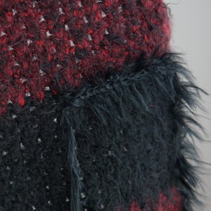 May include: A textured knit garment featuring horizontal stripes of deep red and black yarn. The fabric has a fuzzy, high-pile appearance with visible loose fibres. A prominent black patch pocket with a long, shaggy fringe trim is attached.