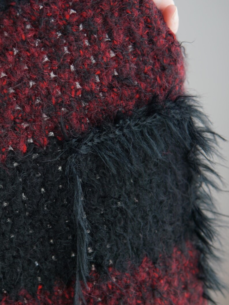 May include: A textured knit garment featuring horizontal stripes of deep red and black yarn. The fabric has a fuzzy, high-pile appearance with visible loose fibres. A prominent black patch pocket with a long, shaggy fringe trim is attached.
