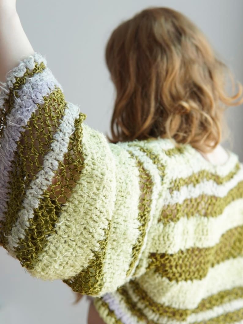 May include: A long-sleeved crochet jumper featuring horizontal stripes in shades of olive green, pale yellow, and light lavender. The garment is constructed with a combination of dense, textured stitches and open, airy mesh sections throughout.