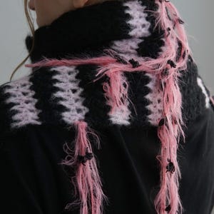 May include: A hand-knitted scarf featuring alternating horizontal stripes of black and light pink yarn. The scarf has a fuzzy, textured appearance with long, loose pink fringe strands hanging down, accented with small black knots throughout the fringe.