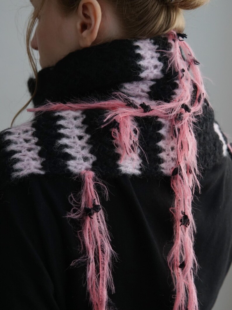 May include: A hand-knitted scarf featuring alternating horizontal stripes of black and light pink yarn. The scarf has a fuzzy, textured appearance with long, loose pink fringe strands hanging down, accented with small black knots throughout the fringe.