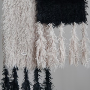 May include: A long, rectangular wall hanging featuring two distinct sections of shaggy, faux-fur-like material in beige and black. The beige section has black tassels at the bottom, while the black section has beige tassels hanging from its base.