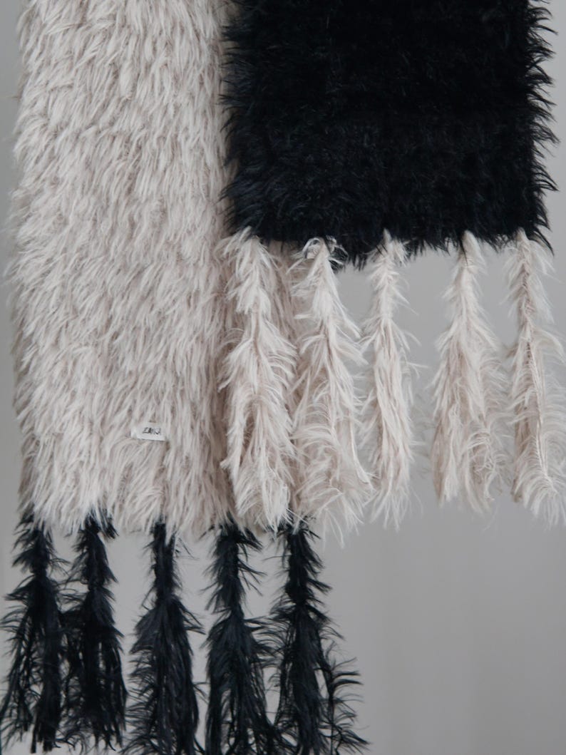 May include: A long, rectangular wall hanging featuring two distinct sections of shaggy, faux-fur-like material in beige and black. The beige section has black tassels at the bottom, while the black section has beige tassels hanging from its base.
