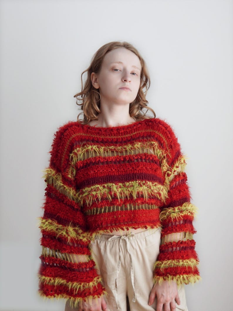 May include: A long-sleeved, cropped sweater featuring a horizontal striped pattern in shades of red, burgundy, and yellow. The garment is constructed with a combination of solid crochet stitches and textured, fuzzy yarn sections, creating an open-knit effect.