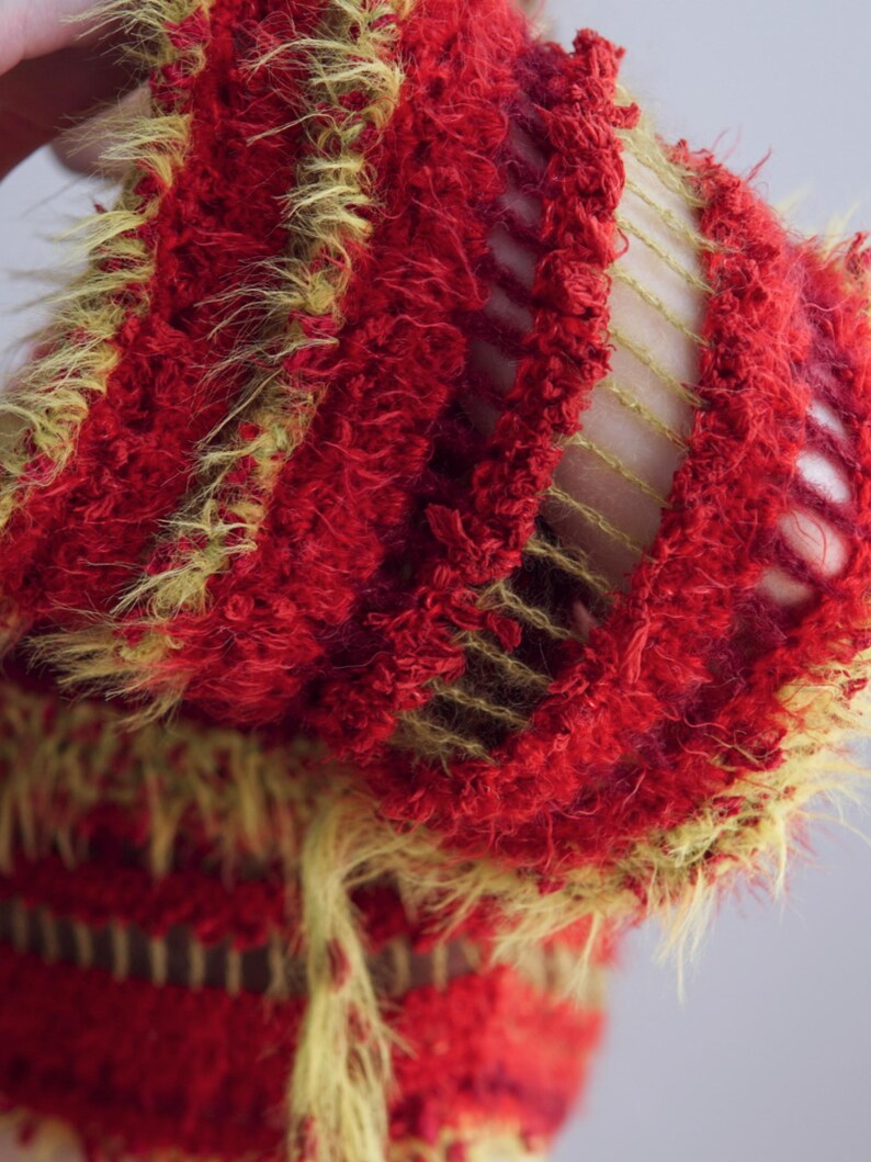 May include: A textured knit garment featuring alternating horizontal bands of fuzzy red yarn and open-work yellow yarn. The fabric displays a distinct contrast between the dense, shaggy red sections and the airy, ladder-like yellow knit structure.
