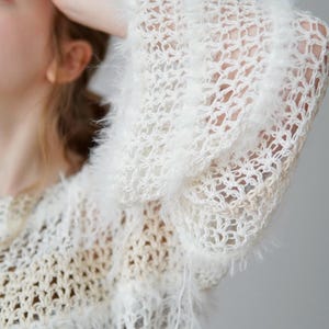 May include: A white, open-knit jumper featuring a loose, airy crochet pattern with integrated fuzzy, textured yarn strips. The garment has long, wide sleeves and a semi-sheer construction, showcasing a layered, multi-dimensional knit design.