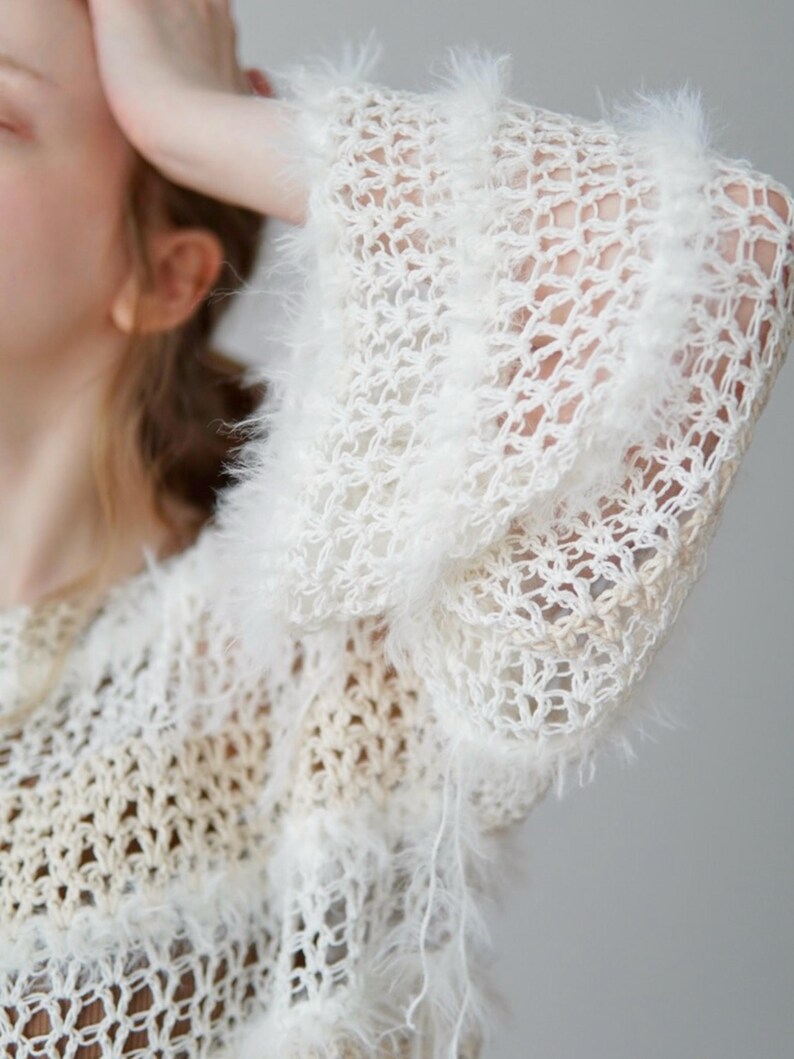 May include: A white, open-knit jumper featuring a loose, airy crochet pattern with integrated fuzzy, textured yarn strips. The garment has long, wide sleeves and a semi-sheer construction, showcasing a layered, multi-dimensional knit design.