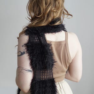 May include: A long, black scarf featuring a combination of open-knit mesh sections and dense, fuzzy faux fur panels. The scarf is draped vertically, showcasing the contrast between the delicate, grid-like structure and the thick, textured black fibers.