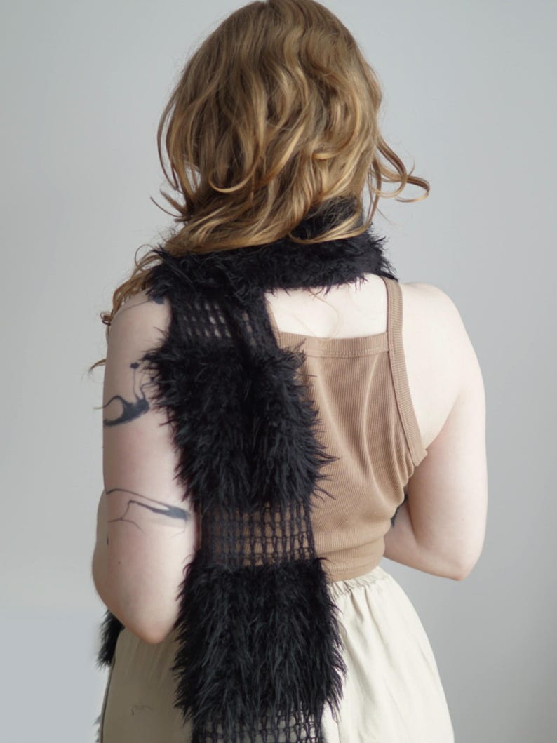 May include: A long, black scarf featuring a combination of open-knit mesh sections and dense, fuzzy faux fur panels. The scarf is draped vertically, showcasing the contrast between the delicate, grid-like structure and the thick, textured black fibers.