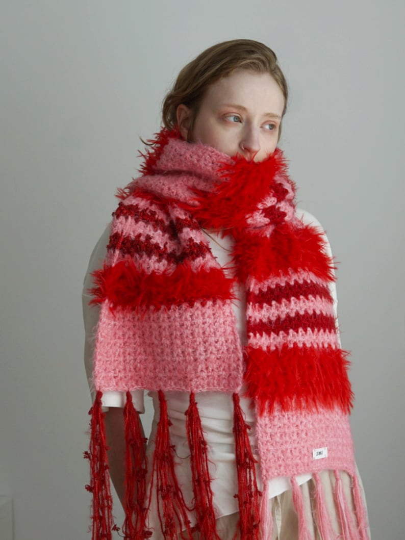 May include: A chunky knit scarf featuring horizontal stripes in shades of light pink, dark pink, and vibrant red. The scarf incorporates textured, fuzzy yarn sections and long, thin red fringe tassels at both ends, with a small white label attached.