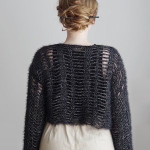 May include: A cropped, long-sleeved sweater in a dark charcoal gray, fuzzy yarn. The garment features a wide, open-knit ladder stitch pattern throughout the back and sleeves, revealing a solid black layer underneath, paired with light beige trousers.