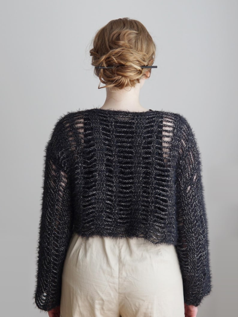 May include: A cropped, long-sleeved sweater in a dark charcoal gray, fuzzy yarn. The garment features a wide, open-knit ladder stitch pattern throughout the back and sleeves, revealing a solid black layer underneath, paired with light beige trousers.