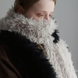 May include: A long, textured scarf featuring a mix of long-pile faux fur in shades of light gray and solid black. The scarf is wrapped around the neck, displaying a shaggy, voluminous appearance with contrasting color blocks of light and dark fibers.