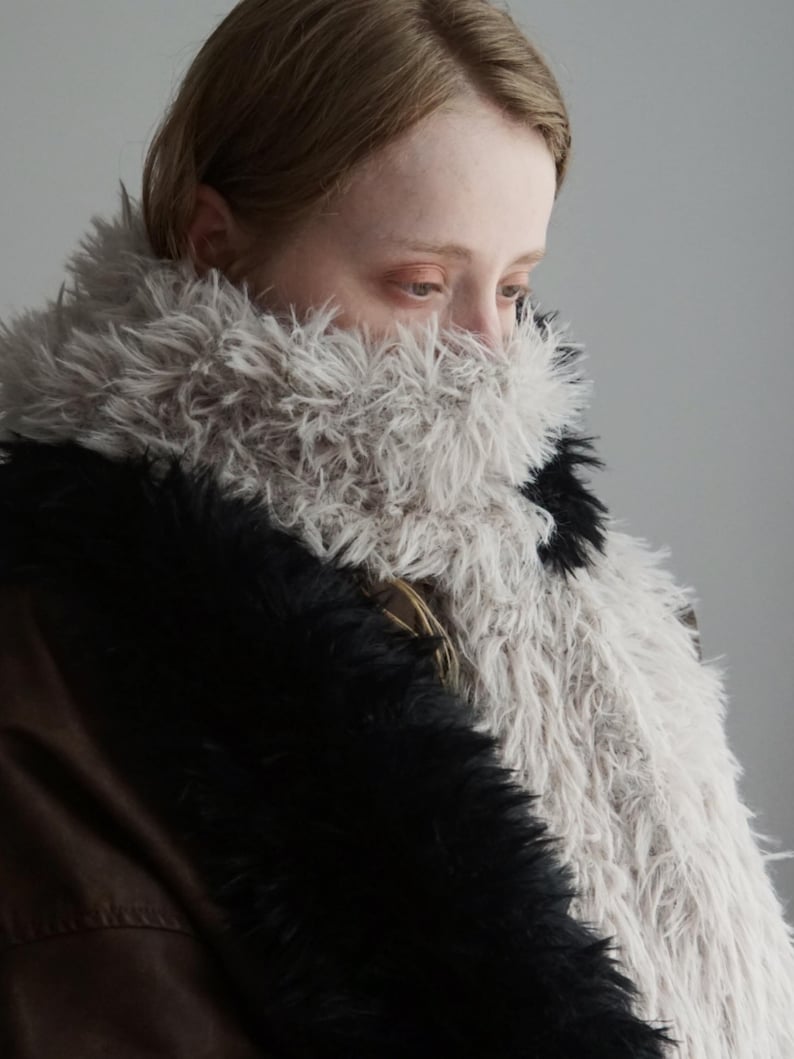 May include: A long, textured scarf featuring a mix of long-pile faux fur in shades of light gray and solid black. The scarf is wrapped around the neck, displaying a shaggy, voluminous appearance with contrasting color blocks of light and dark fibers.