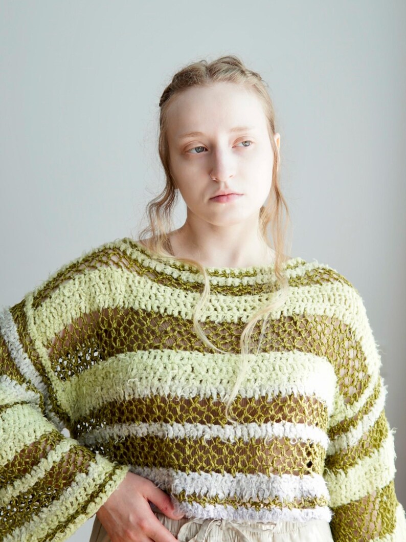 May include: A long-sleeved, loose-knit jumper featuring horizontal stripes in shades of light lime green, olive green, and white. The garment is constructed with a combination of textured, fuzzy yarn and an open, airy crochet stitch pattern.