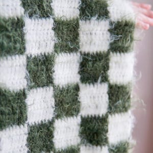 May include: A crocheted blanket featuring a checkered pattern of alternating dark olive green and cream white squares. The green squares are crafted from a fuzzy, textured yarn, while the white squares consist of a smooth, standard yarn.
