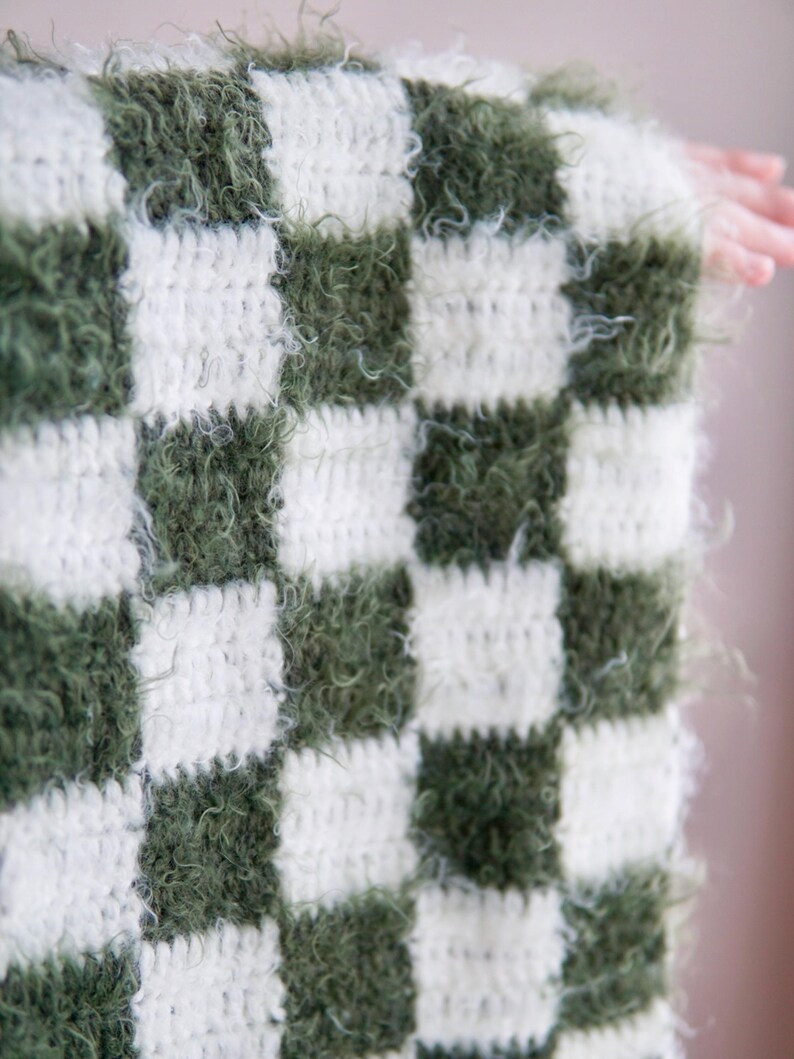May include: A crocheted blanket featuring a checkered pattern of alternating dark olive green and cream white squares. The green squares are crafted from a fuzzy, textured yarn, while the white squares consist of a smooth, standard yarn.
