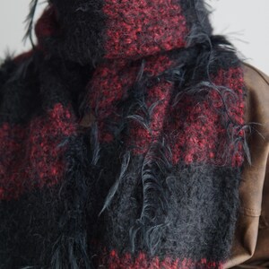 May include: A chunky knit scarf featuring a bold plaid pattern in deep black and dark red. The fabric has a fuzzy, textured appearance with loose, frayed yarn strands hanging from the surface, creating a distressed look around the neck.