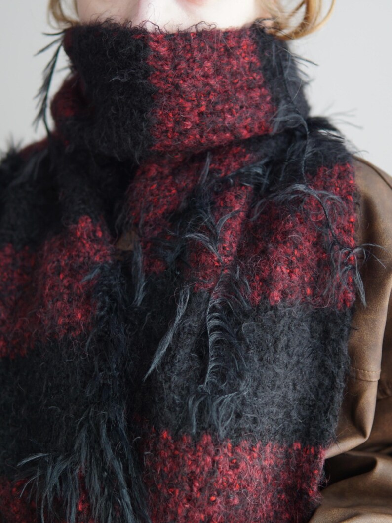 May include: A chunky knit scarf featuring a bold plaid pattern in deep black and dark red. The fabric has a fuzzy, textured appearance with loose, frayed yarn strands hanging from the surface, creating a distressed look around the neck.