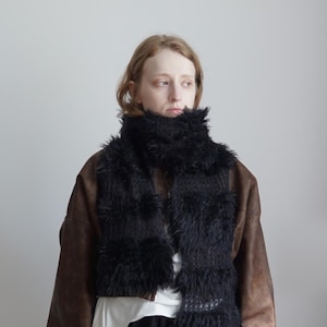 May include: A black scarf featuring alternating sections of long, shaggy faux fur and a textured, open-knit crochet pattern. The scarf is wrapped around the neck and hangs down the front, displaying a small white rectangular label at the bottom.