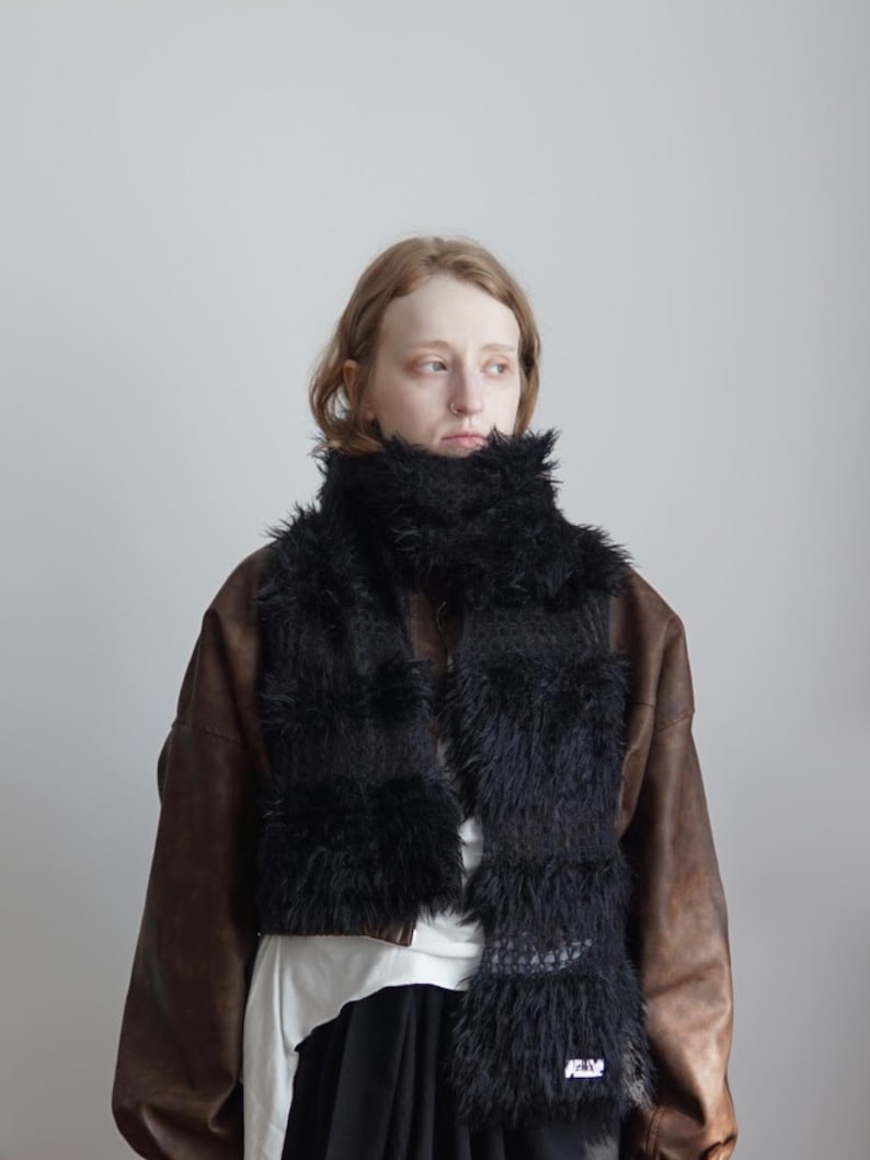 May include: A black scarf featuring alternating sections of long, shaggy faux fur and a textured, open-knit crochet pattern. The scarf is wrapped around the neck and hangs down the front, displaying a small white rectangular label at the bottom.