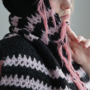 May include: A hooded garment featuring a horizontal striped pattern in black and light pink. The knit fabric has a fuzzy, textured appearance with loose, frayed pink yarn strands hanging from the edges of the hood and the front opening.
