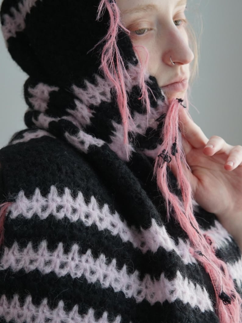 May include: A hooded garment featuring a horizontal striped pattern in black and light pink. The knit fabric has a fuzzy, textured appearance with loose, frayed pink yarn strands hanging from the edges of the hood and the front opening.