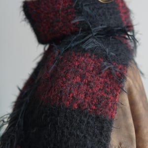 May include: A thick, fuzzy scarf featuring a bold, alternating block pattern of deep black and dark red. The knit texture is highly visible, with loose, wispy fibres extending from the surface, creating a soft, voluminous appearance around the neck.