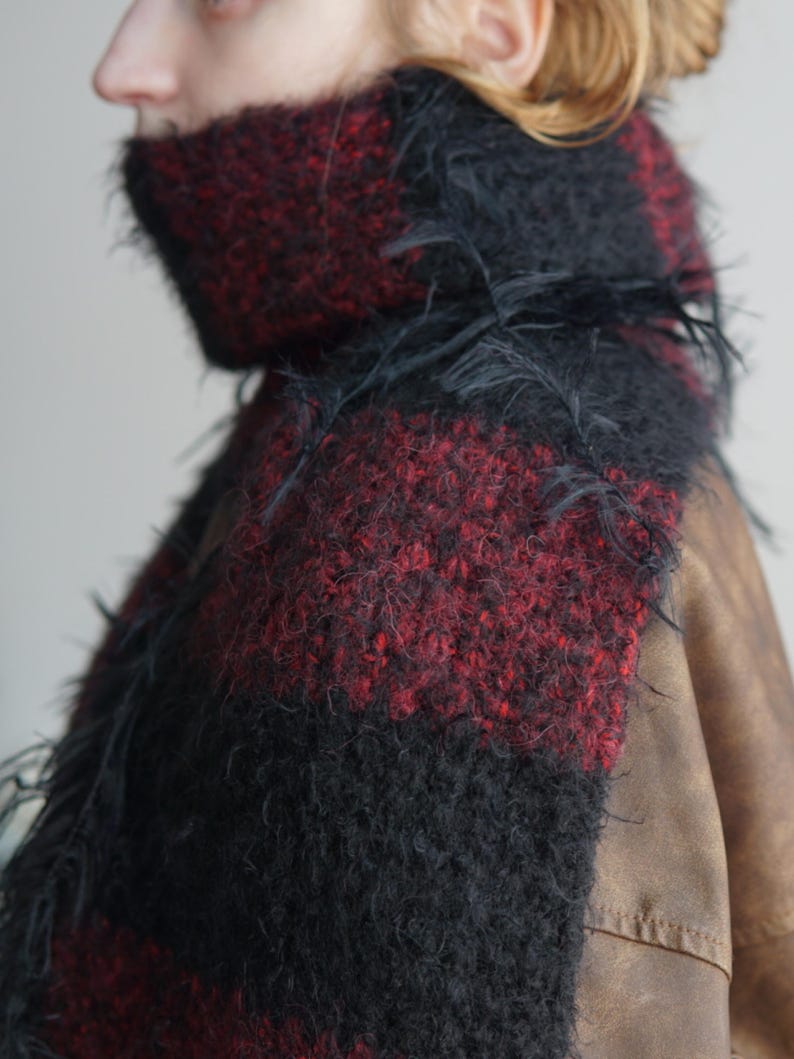 May include: A thick, fuzzy scarf featuring a bold, alternating block pattern of deep black and dark red. The knit texture is highly visible, with loose, wispy fibres extending from the surface, creating a soft, voluminous appearance around the neck.