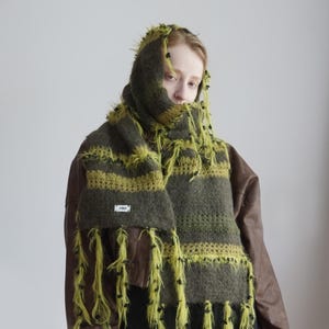 Op de afbeelding: Een pluizige, oversized sjaal met horizontale strepen in olijfgroen en limoengroen, geaccentueerd met lange, ruige limoengroene franjes en kleine zwarte knoopjes. De sjaal is voorzien van een klein wit rechthoekig label met zwarte tekst.