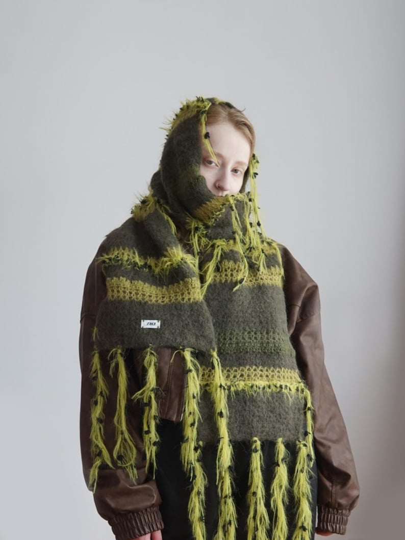 Op de afbeelding: Een pluizige, oversized sjaal met horizontale strepen in olijfgroen en limoengroen, geaccentueerd met lange, ruige limoengroene franjes en kleine zwarte knoopjes. De sjaal is voorzien van een klein wit rechthoekig label met zwarte tekst.