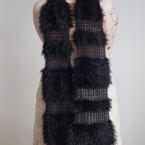 May include: A long black scarf featuring alternating horizontal bands of open-knit mesh fabric and thick, shaggy faux fur. The scarf has a rectangular shape with a consistent width throughout, showcasing a textured contrast between the smooth mesh sections and the dense, fuzzy fur panels.