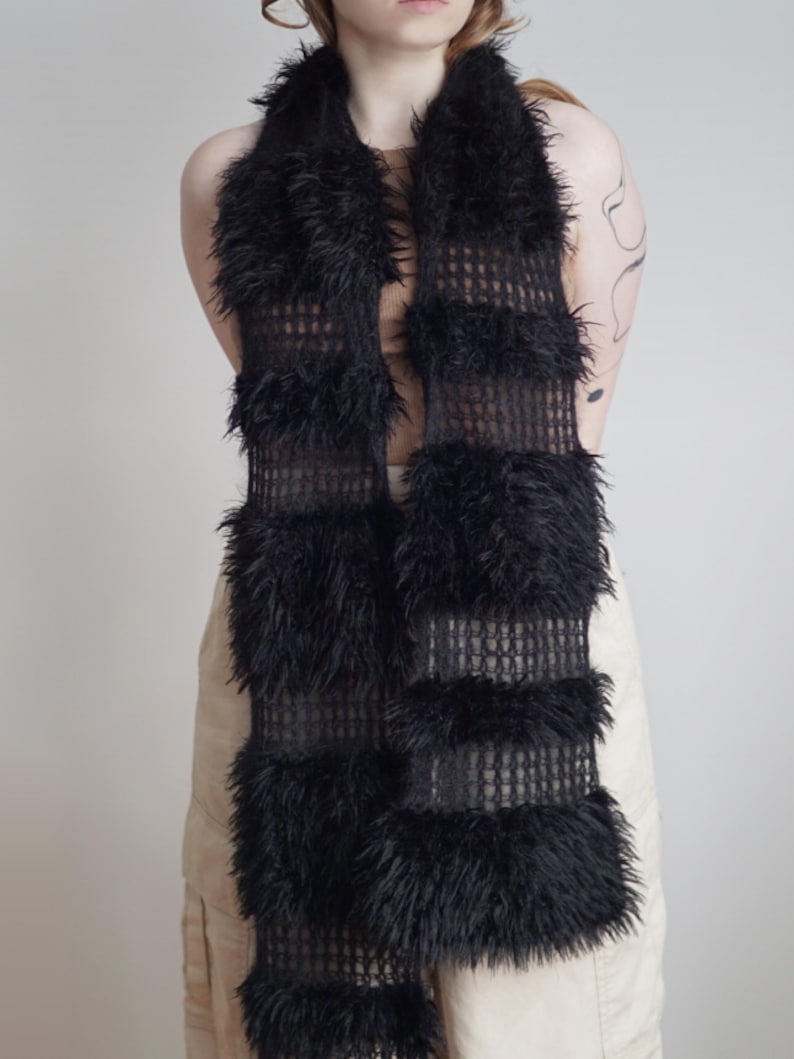 May include: A long black scarf featuring alternating horizontal bands of open-knit mesh fabric and thick, shaggy faux fur. The scarf has a rectangular shape with a consistent width throughout, showcasing a textured contrast between the smooth mesh sections and the dense, fuzzy fur panels.