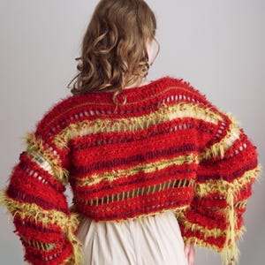 May include: A cropped, long-sleeved sweater featuring a textured, shaggy knit pattern in vibrant shades of red, burgundy, and lime green. The garment includes horizontal open-knit stripes and loose, frayed yarn edges throughout the design.