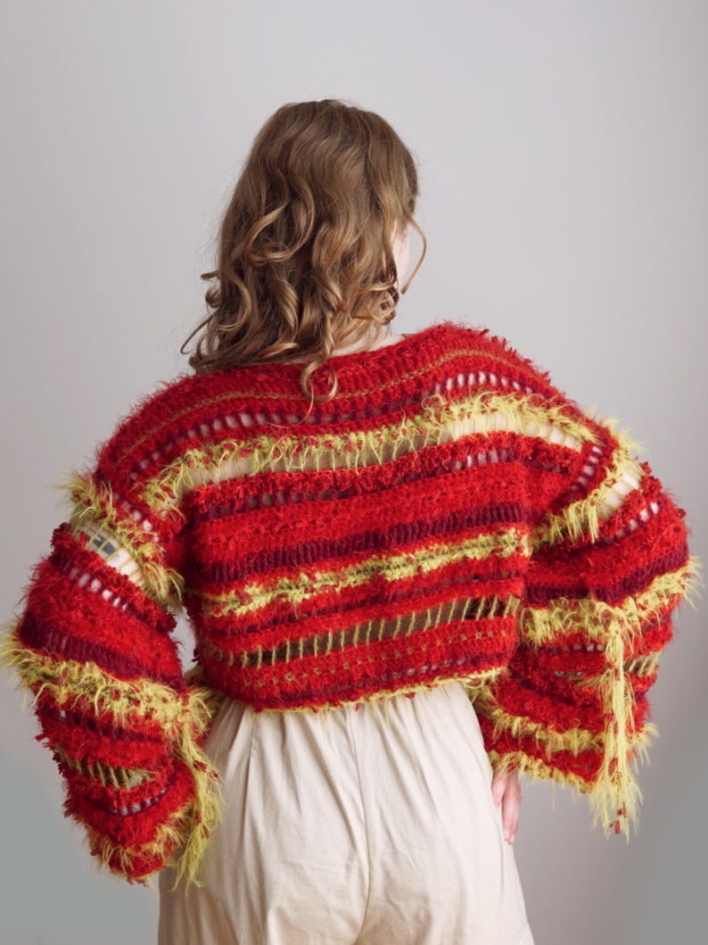 May include: A cropped, long-sleeved sweater featuring a textured, shaggy knit pattern in vibrant shades of red, burgundy, and lime green. The garment includes horizontal open-knit stripes and loose, frayed yarn edges throughout the design.