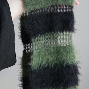 May include: A fuzzy scarf featuring alternating horizontal stripes of olive green and black. The design incorporates sections of dense, textured yarn alternating with bands of a more open, sheer, and grid-like knit pattern throughout the length.