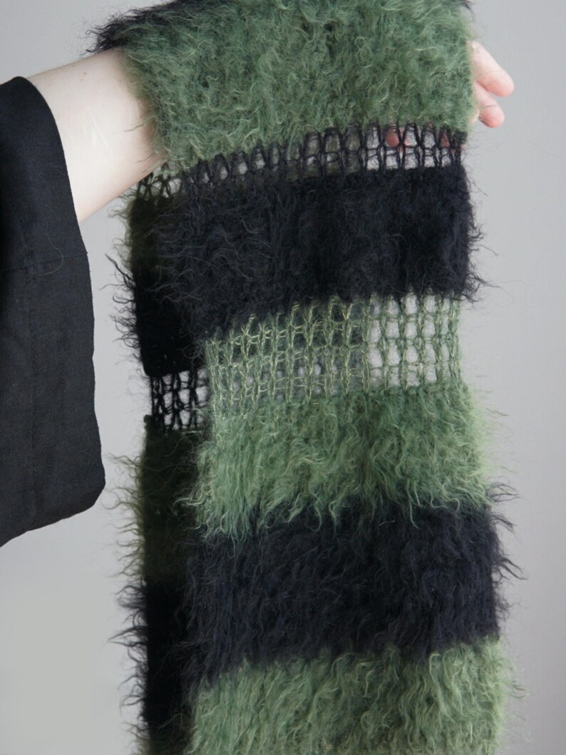 May include: A fuzzy scarf featuring alternating horizontal stripes of olive green and black. The design incorporates sections of dense, textured yarn alternating with bands of a more open, sheer, and grid-like knit pattern throughout the length.