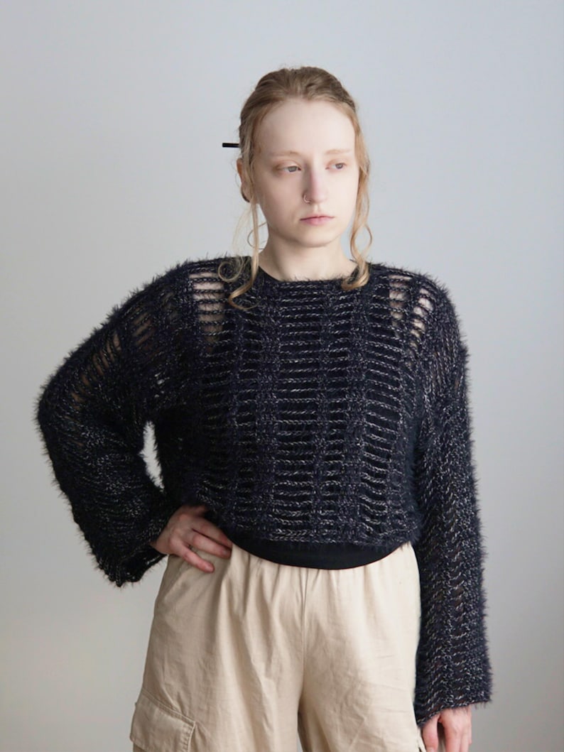 May include: A black fuzzy long-sleeve sweater with a horizontal ladder-stitch pattern and a wide crew neckline. The knit fabric has a textured, hairy appearance with subtle silver metallic threads woven throughout the dark yarn.