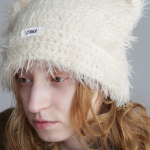 May include: A cream-colored, fuzzy, faux-fur beanie with two pointed ear-like shapes at the top. The hat features a white rectangular fabric label on the front with the word ZAIKA printed in black text. The texture is long, shaggy, and soft.