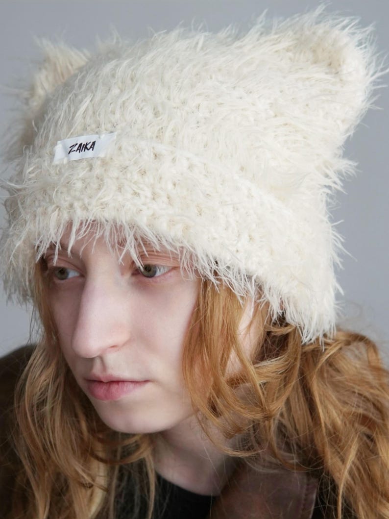 May include: A cream-colored, fuzzy, faux-fur beanie with two pointed ear-like shapes at the top. The hat features a white rectangular fabric label on the front with the word ZAIKA printed in black text. The texture is long, shaggy, and soft.