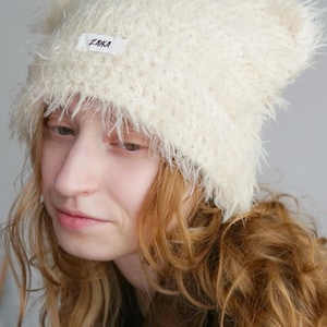 May include: A cream-colored, fuzzy, textured beanie with two pointed ear-like shapes at the top. A small, rectangular white fabric label is sewn onto the front center of the beanie, featuring the black text ZAIKA printed in a simple font.