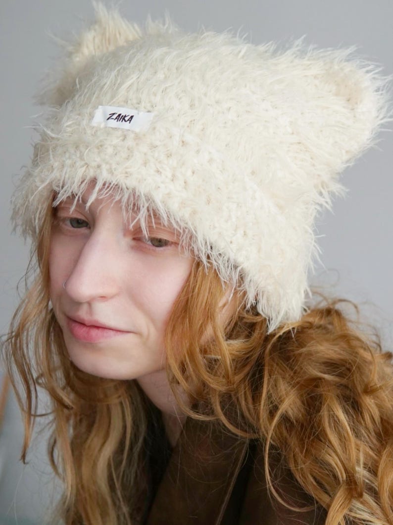 May include: A cream-colored, fuzzy, textured beanie with two pointed ear-like shapes at the top. A small, rectangular white fabric label is sewn onto the front center of the beanie, featuring the black text ZAIKA printed in a simple font.