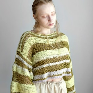 May include: A cropped, long-sleeved jumper featuring a loose, open-knit crochet construction. The garment displays horizontal stripes in alternating shades of olive green, pale yellow, and light lavender, with a wide, rounded neckline.