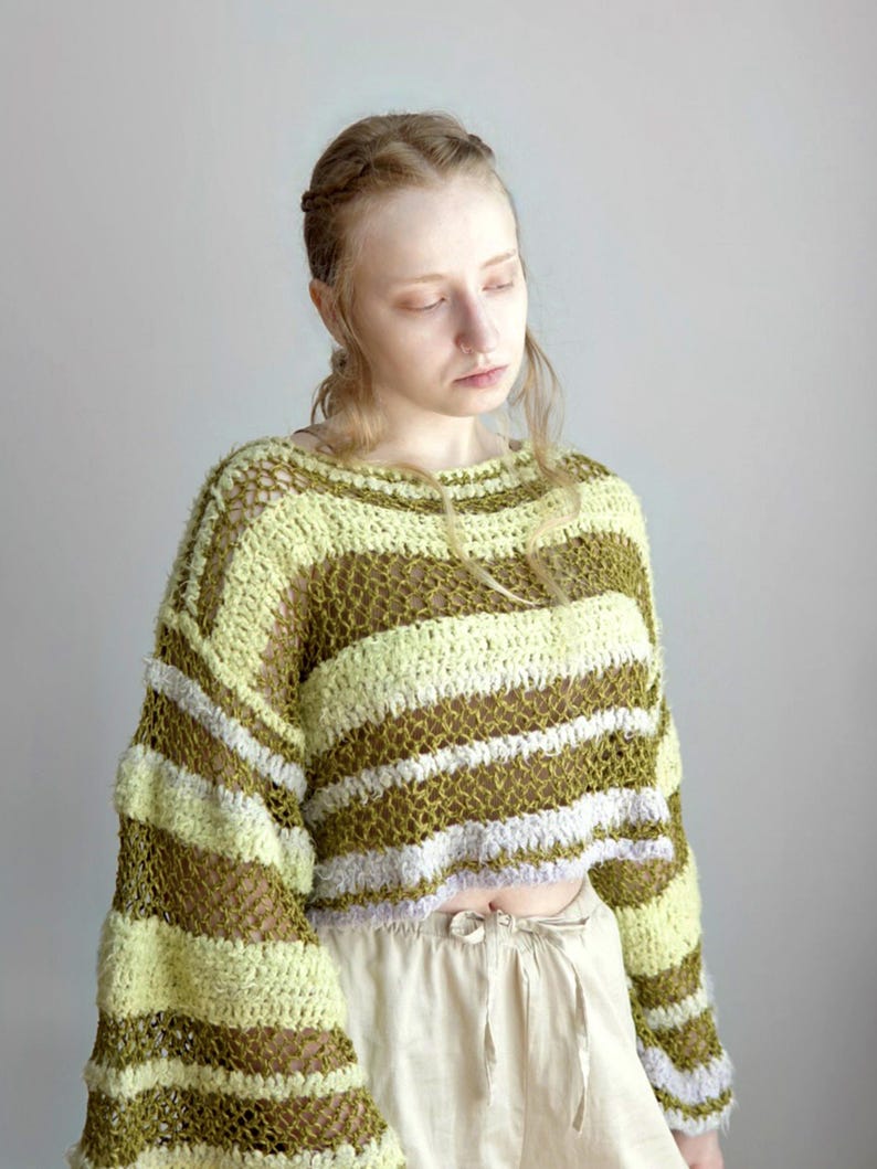 May include: A cropped, long-sleeved jumper featuring a loose, open-knit crochet construction. The garment displays horizontal stripes in alternating shades of olive green, pale yellow, and light lavender, with a wide, rounded neckline.